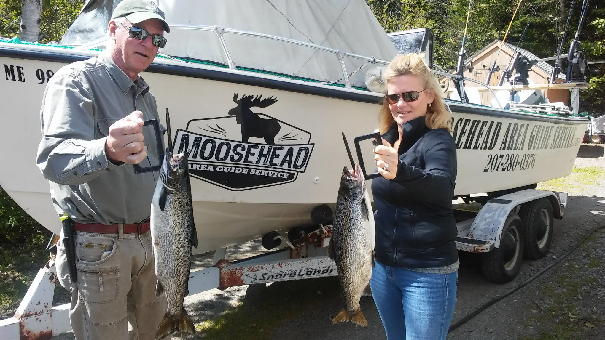 A couple standing in front of boat, each holding a landlocked salmon they caught on a Greenville, Maine Fishing Charter